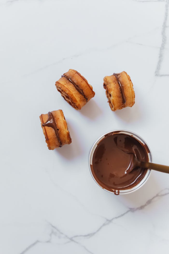 Three chocolate drizzled pastries and a bowl of melted chocolate on marble surface.